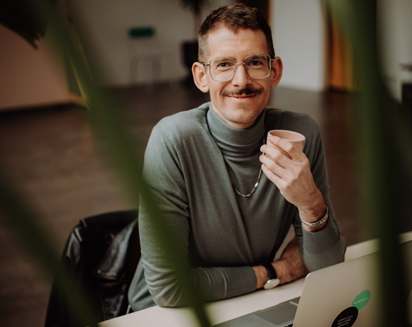 Benni Bauerdick unser Experte für Sprechen Stimme und Souveränes Auftreten sitzt an einem Schreibtisch mit einem Kaffee in der Hand und schaut in die Kamera