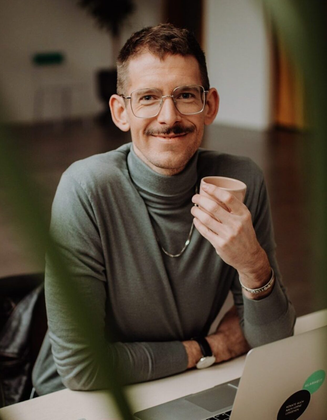 Benni Bauerdick unser Experte für Sprechen Stimme und Souveränes Auftreten sitzt an einem Schreibtisch mit einem Kaffee in der Hand und schaut in die Kamera
