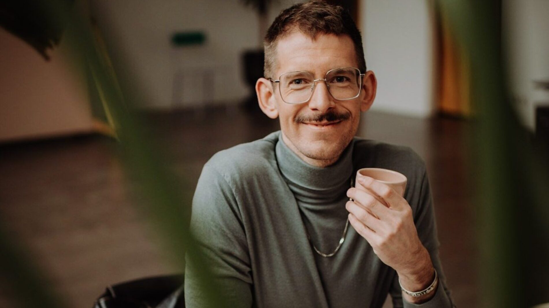 Benni Bauerdick unser Experte für Sprechen Stimme und Souveränes Auftreten sitzt an einem Schreibtisch mit einem Kaffee in der Hand und schaut in die Kamera