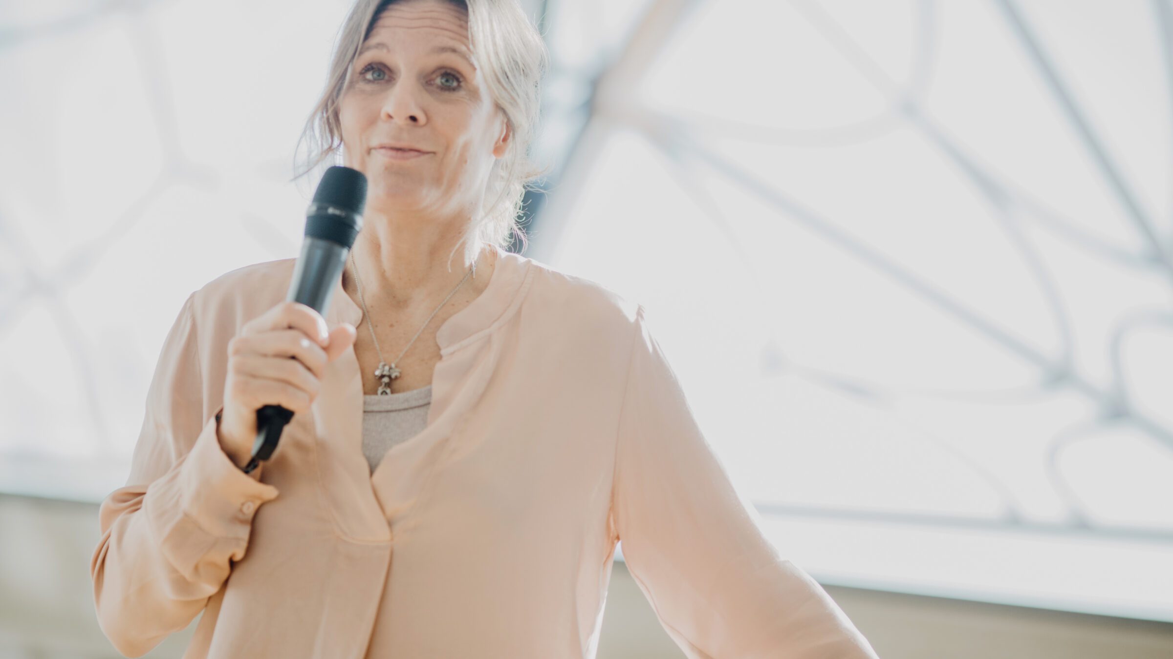 Trauerrednerin Tina Bartels in heller Kleidung mit Mikrofon hält eine Rede vor dem großen prunkvollen Fenster.
